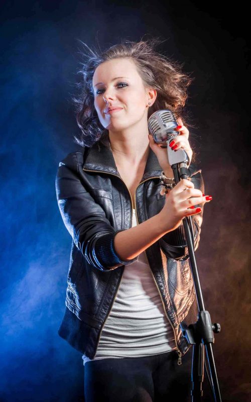 Cantante interpretando una emotiva canción en un concurso internacional de canto, con luces escénicas y un público entusiasta en el fondo